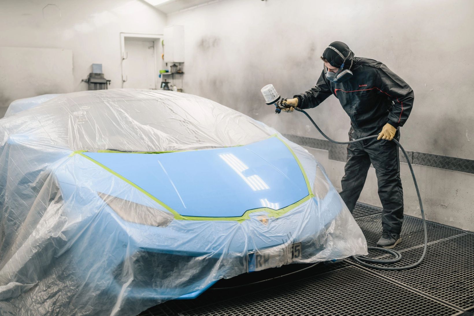 Lackierer in Schutzkleidung lackiert in der Lackierkabine ein abgeklebtes Auto